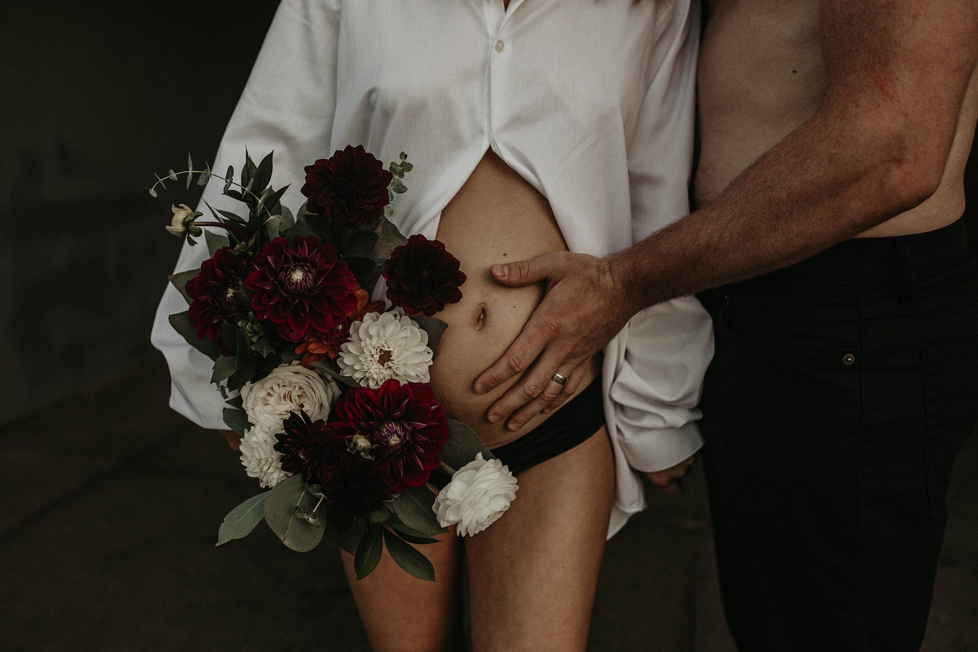 Pregnant couple holds a floral bouquet on the shore of the beach.